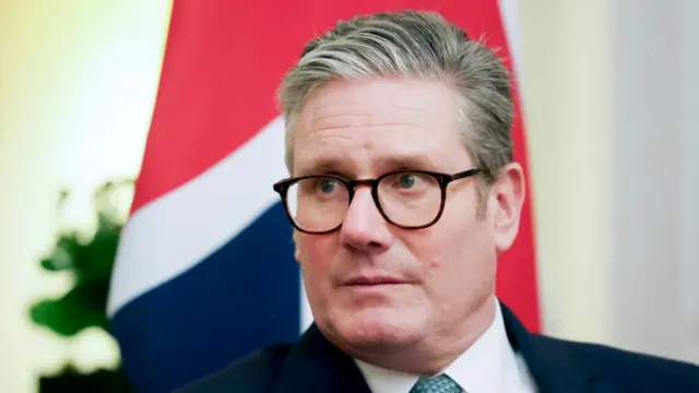 Close-up shot of Keir Starmer sitting in front of a Union flag. He's wearing a black suit with the top of a patterned blue visible at the bottom centre of the image