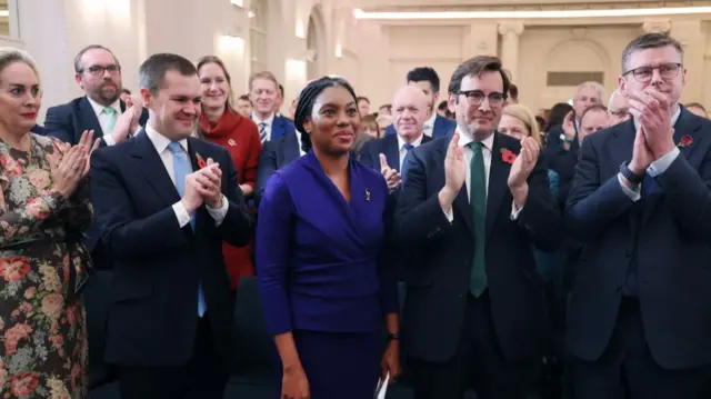 Robert Jenrick (L) applauds Kemi Badenoch as she's elected. Badenoch is at the centre of the image smiling, her husband Hamish in dark suit and green tie cheering next to her