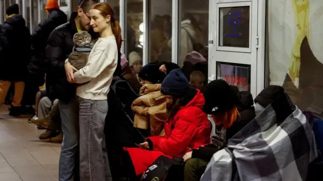 People wearing beanies and blankets sit on a tiled floor. Nearby, a woman holds a cat in her arms