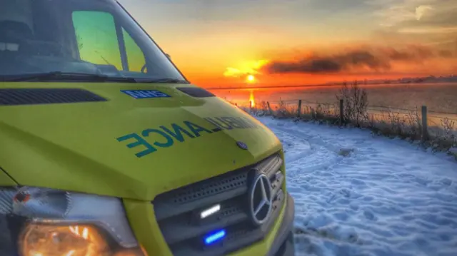 A yellow ambulance parked in snow beside a field with a fence surrounding it and the sunrise in the background