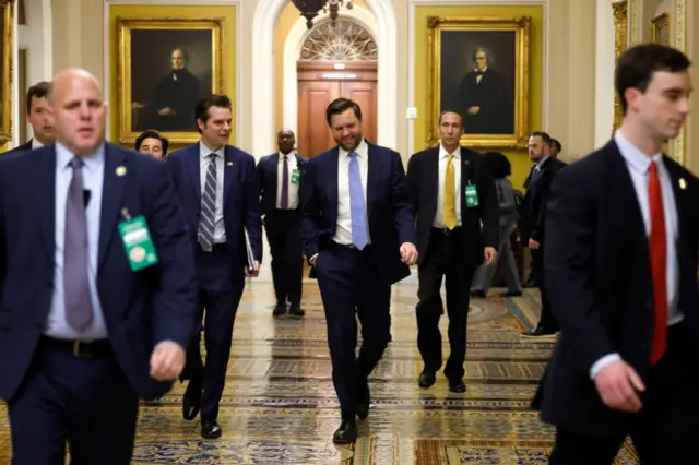 Men in suits flank JD Vance who walks down a tiled hallway with his hand in his right pocket