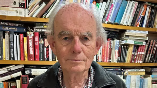 A balding man with white hair stands in front of bookshelves stuffed with books about famous political figures. He is wearing a checked shirt and fleece and looking directly at the camera.