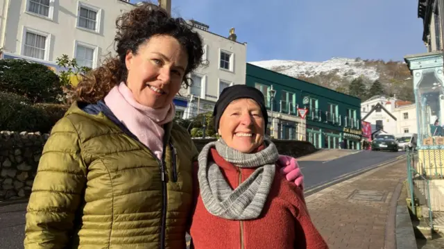 Two women, one in a green coat and the other in a red coat, both wearing hats and scarves, standing in a high street with snow on the hills behind them