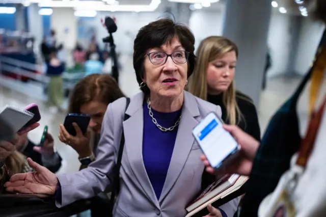 Woman wearing glasses and grey jacket is surrounded by reporters asking her questions