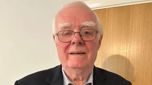 A man with white hair and wire-frame glasses. He is wearing a shirt and black suit jacket and standing in front of a wall and wooden door. He has white hair and eyebrows. His right eye is partially closed.