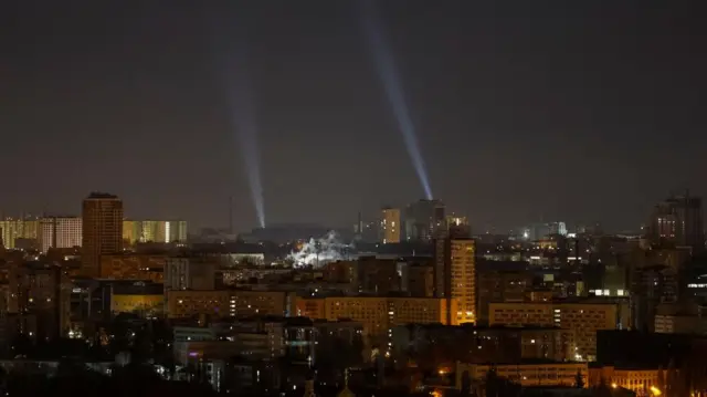 A view of Kyiv at night, with city buildings illuminated and two spotlights drawing two streaks of light through the sky