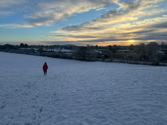 The sun rising above rooftops in Shrewsbury. A child in a red jacket stands in the middle of a snow-covered field looking towards trees and a railway line