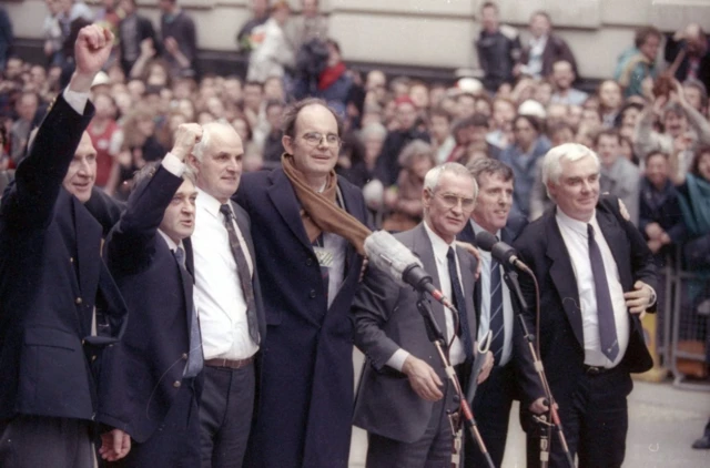 Six men in suits stand in front of microphones on a street, with a crowd gathered behind them. The two men on the right have their right fist in the air. All the men are smiling.