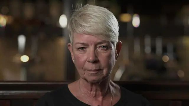 A woman with short blond hair swept across her forehead. She is wearing a black top and looking into the camera. Behind her but out-of-focus is a pub backdrop.