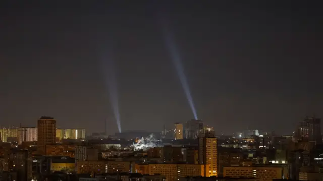 Two beams of light shine in a dark sky in a cityscape