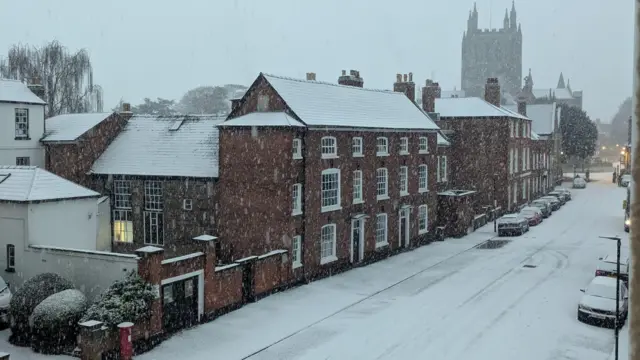 A snowy street scene with historic two and three storey buildings facing on to a whitened carriageway with snow-covered cars parked along it.