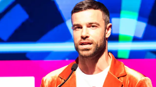 Ben Starr speaking on stage, wearing an orange jacket