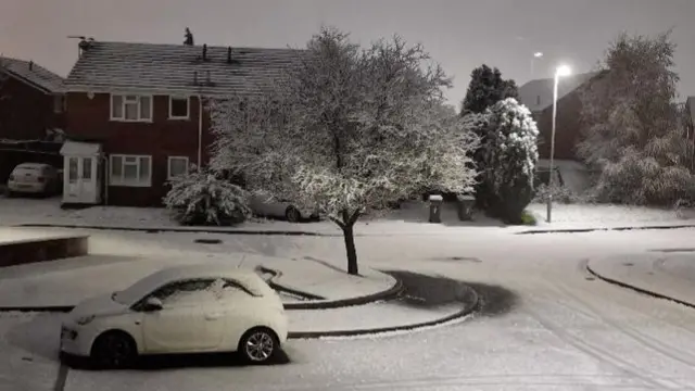 A street covered in snow with a white car in the foreground an a house surrounded by trees in the background