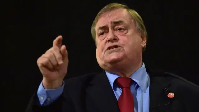 John Prescott mid-shot cutting off below the shoulders. He's wearing a black suit, blue shirt and red tie, his right hand extended and his pointer finger up