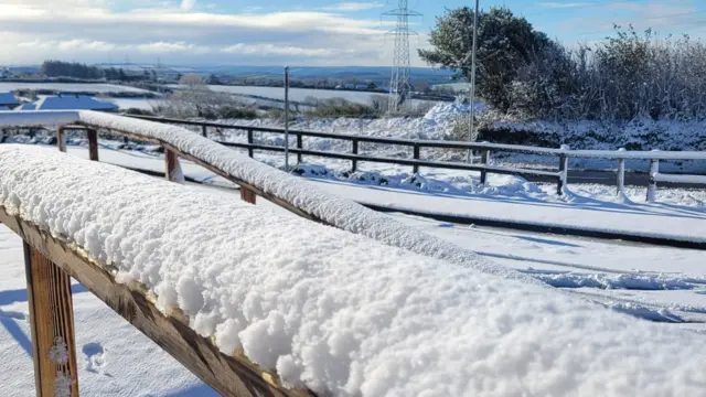 Deep snow covers wooden handrails in the foreground and the countryside in the distance is covered in snow.