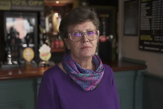 A woman in a purple jumper and snakeskin pattern purple turquoise and pink scarf. She is wearing glasses and has short brown hair. A bar counter and beer taps and bottles can be seen behind her.