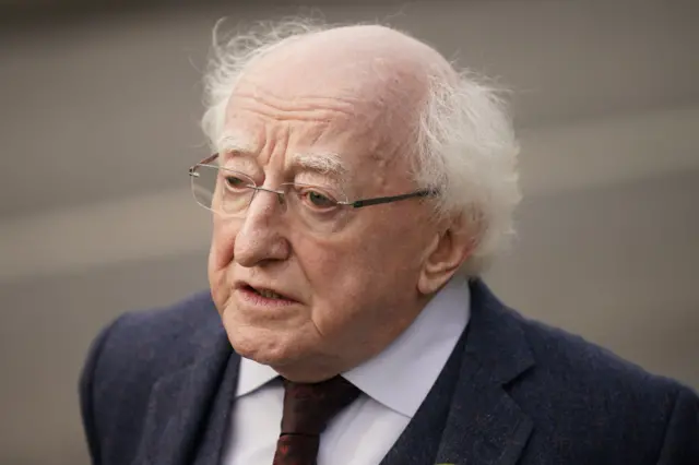 Michael D Higgins. He is balding, with white hair, rimless glasses. He is wearing a dark suit, white shirt and plum tie.