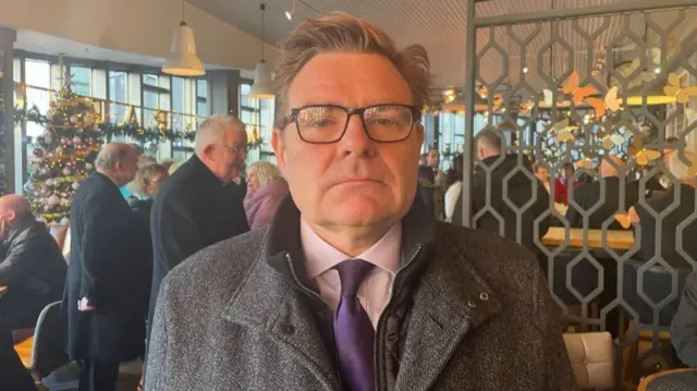 A man in a grey flecked coat, with a pink shirt and purple tie. He is wearing glasses and has light brown hair. he is standing in a cafe. In the background other men in dark coats can be seen chatting. There are festive decorations around the edge of the cafe, which has a wall of windows and pendant lights hanging from the ceiling.