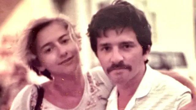 A couple in an old colour photograph. A woman on the left has her arm around a man. She has blond hair and is wearing a white top with applique pattern around the shoulders. The man has dark hair, a moustache, and is wearing an open colllar pale shirt.