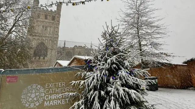 Heavy snow falls on Exeter Christmas Market.Exeter Cathedral is in the background. A  sign states Exeter Cathedral Christmas Market. A tree is covered in snow.