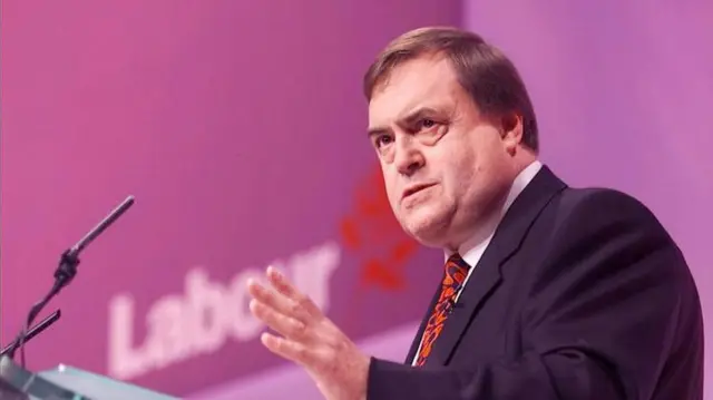 Prescott delivers speech at Labour Party conference in Brighton in 2000. He's in black suit, patterned red/black tie and white suit, a lectern and mic in front of him. He ha his left hand reaised, background is blurred