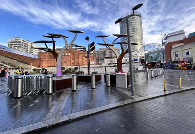 A tree memorial sculpture, featuring three trees made from steel. It is on an outside paved area, with bollards around it and metal barriers lean against it. In the background city high-rise buildings can be seen, including the Odeon and the Rotunda.