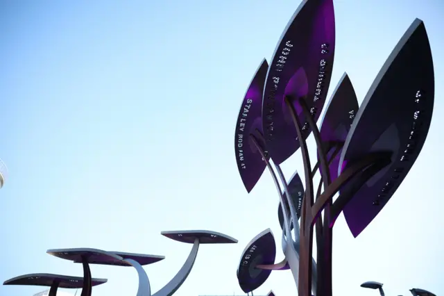 Leaves of a steel tree sculpture memorial, with names of bombing victims Stanley Bodman and John Rowlands visible. A blue sky forms the backdrop.