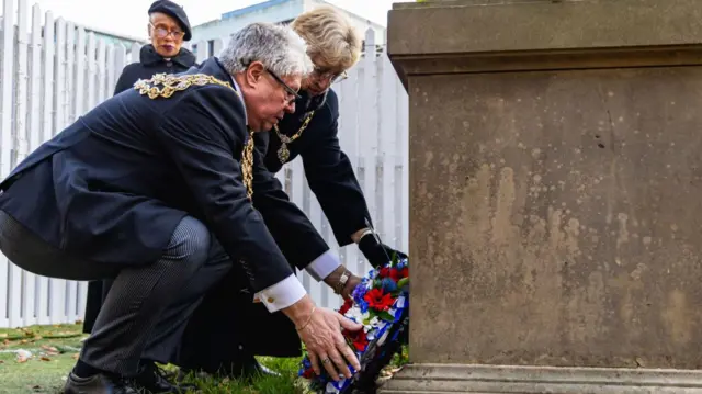 A man and woman crouch at the foot of a stone plinth to lay a wreath of purple, red, yellow and white flowers. Both are dressed in black, with mayoral chains around their necks. In the background a woman dressed in a black coat and hat can also be seen.