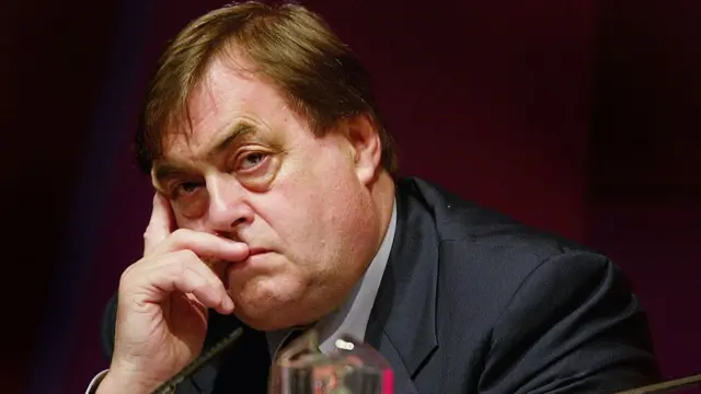 John Prescott looking pensive as he looks to his left, his head leaning on his right had. He's wearing a dark blue suit, white shirt