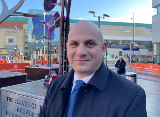 A man  in a black jacket, blue tie and white shirt standing in front of an outdoor steel tree memorial sculpture. He is bald. In the background a shopping centre can be seen.