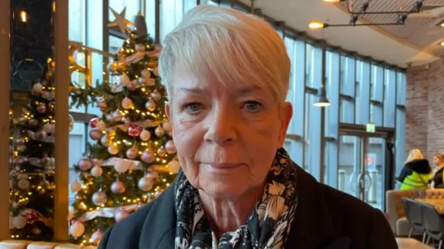 A woman with short blonde hair, in a black jacket and pink, white and black patterned scarf, sits in a cafe. A Christmas tree with pink baubles and lights can be seen in front of a wall of windows in the background.