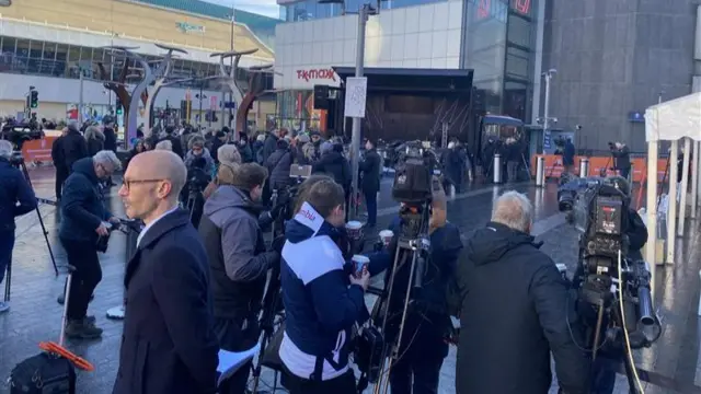 A large group of people, many of them with TV cameras, are gathered in an outdoor area