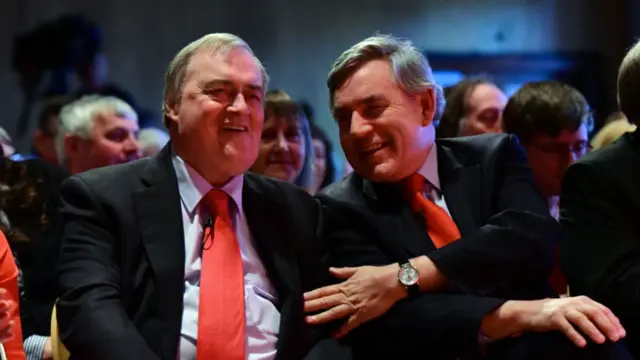 John Prescott (L) smiling as he speaks to Gordon Brown (R). Prescott is sitting down wearing a black suit, blue shirt and red tie. Brown is leaning into Prescot''s left shoulder, his left hand on Prescott's left arm as he smiles