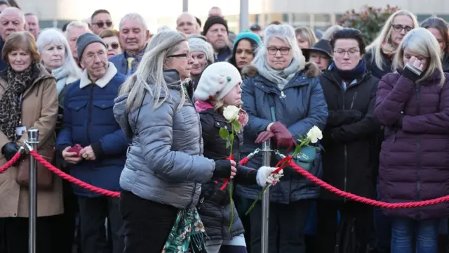 A woman and a child carrying white roses, with people gathered behind a red cordon