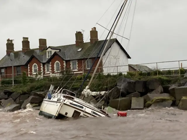 The image shows a boat that appears to be sinking. The vessel is white and blue. Half of the boat is underwater and is pinned up against the shoreline.