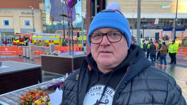 Sully wearing a blue hat a warm coat and a Birmingham pub bombings jumper. There are people in hi-vis behind him and some flowers.