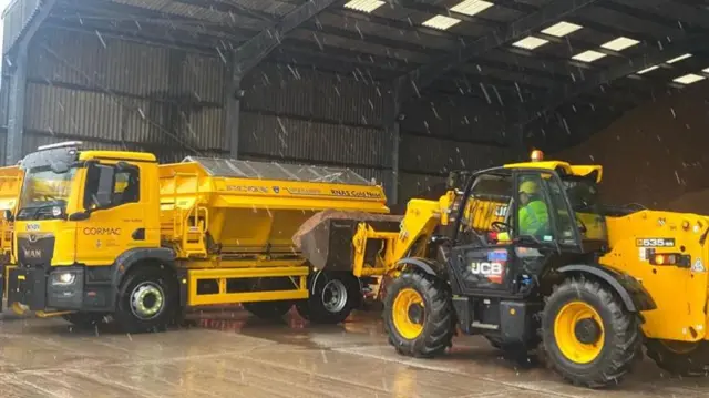 A gritter being loaded with salt in front of a large pile of salt