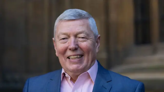 Alan Johnson smiling in close-up shot. He's wearing a dark grey suit, pink shirt with no tie. A blurred stone building is behind him