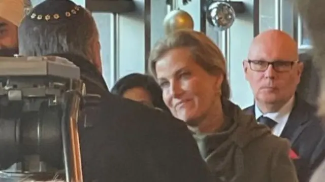 Sophie, Duchess of Edinburgh, chats to a man with his back to the camera. Other guests, including a man in glasses and a suit, stand around her. The Duchess has short blonde hair and is wearing a green coat with a bow on the sholder.