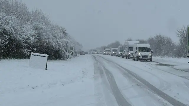 The A37 road covered in snow. There are cars and vans on the road but they are not moving.