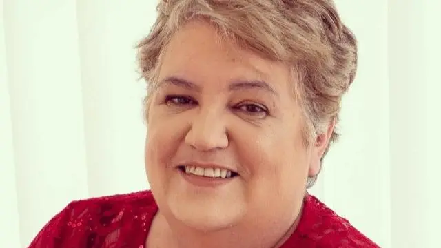 A woman with short light brown hair smiles at the camera. She is wearing a red top and sitting in front of white slatted blinds.