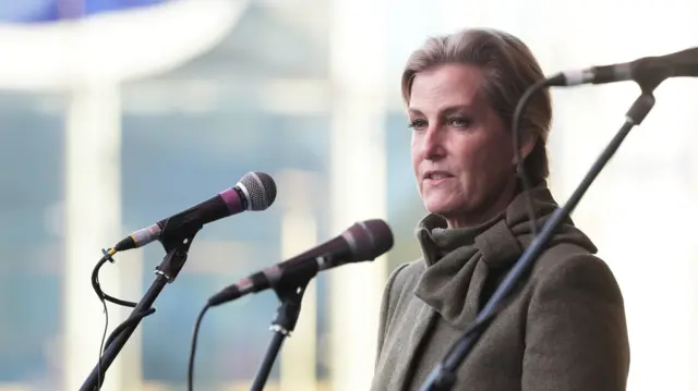 Sophie, Duchess of Edinburgh, gives a speech standing in front of three microphones. She is wearing a green coat