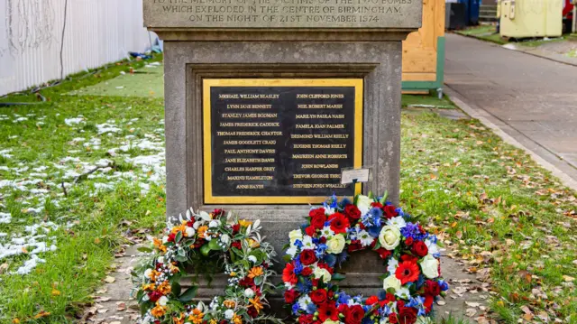 A black memorial plaque framed in gold, with gold writing, on a stone plinth. Two floral wreaths lie at the bottom of it. The wreath on the left is green, orange, white and red. the other is red, purple, yellow and green. Grass surrounds the plinth, with an adjacent path and fencing.