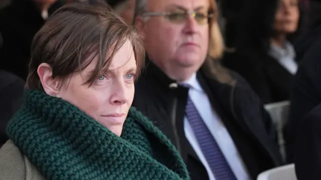 MP Jess Phillips, in a dark green knitted scarf and green coat, looks pensively forward. She is seated on a row, with other guests out of focus next to and behind her. She has short brown hair, swept across her forehead.