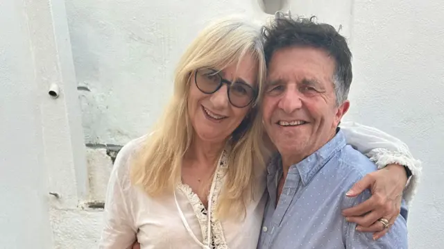 A woman with long blond hair and sunglasses with her arm around a man with dark greying hair, wearing a blue shirt with white spots. She is wearing a white dress and the pair are standing in front of a white wall. Both are smiling.