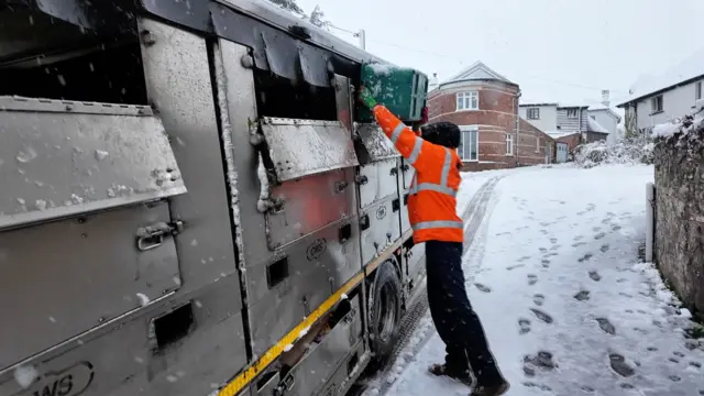 Photo shows a waste collection worker loading up a silver waste collection vehicle. The ground is covered in snow and flurries are still coming down.