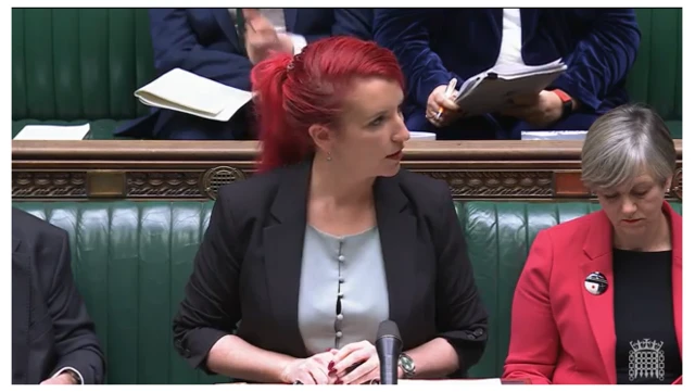 Louise Haigh stands at the dispatch box in grey silk blouse wiht buttons to the front and black jacket, her hair in a ponytail. Labour MP Lilian Greenwood sits on the benches to her left