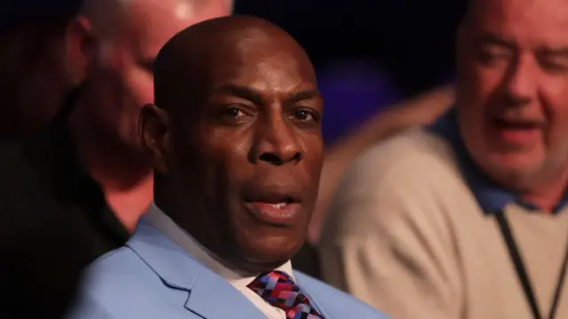 Frank Bruno sitting ringside prior to the Lightweight fight. He's wearing a powder blie suit, patterned tie and white shirt. A blurred man can be seen sitting to his left