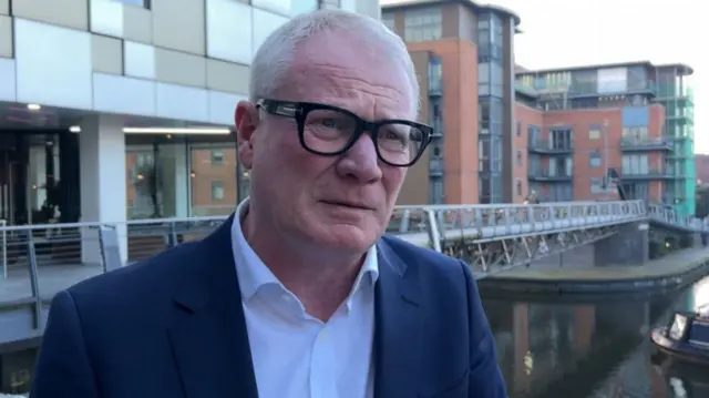 A man with grey hair, dark framed glasses and a navy suit jacket and white shirt. He is standing in front of a canal, with low-rise buildings along its bank.