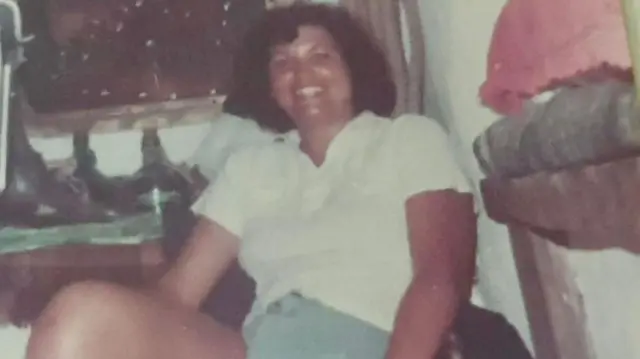 A faded photo of woman in a white t-shirt and blue shorts with short brown hair, smiling at the camera. She is reclining on a seat in a room with various bottles and cushions around her.